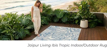 A woman stands on a wooden deck next to a cream rug with blue tropical leaves. Image Text: Jaipur Living Ibis Tropic Indoor/Outdoor Rug