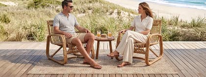 A man and woman face each other in rattan armchairs on a tan rug.
