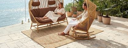 A couple sits on matching woven canopy furniture over a tan rug on a terrace.