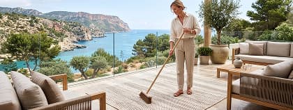 A woman sweeps a neutral patterned area rug on a balcony overlooking a scenic coastline.