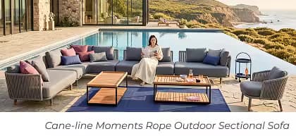 Woman reads on a large modular outdoor sectional with teak coffee tables beside an infinity pool and ocean.